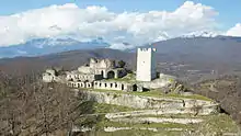 Photograph of a ruined medieval stone-built fortress on a green hilltop in autumn, with the Caucasus mountains in the background