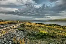 A picture of Rybachy Peninsula on a partly cloudy July day. The Soviet Union demanded that Finland cede the peninsula, its northernmost point at the time, along with other areas. The sun is either rising or setting and shines on the long grass at an angle. A body of water, the Barents Sea, fills a quarter of the picture. A gravel road leads to a lone house in the distance.
