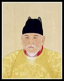 An old aged bearded man wears yellow robes with dragons inscribed and a black hat.