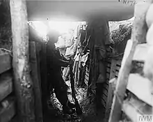 A member of the 1/7KR, stands in silhouette, while leaning on the wooden wall of a covered portion of a trench. Two additional soldiers stand in the background, in an uncovered part of the trench, almost washed out by the contrast.