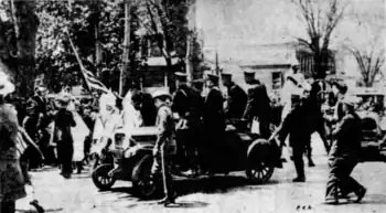 A black-and-white photograph showing several hooded Ku Klux Klan members, one of whom is carrying an American flag. A few policemen are near the Klansmen, and more officers stand in a nearby car. Civilians are intermixed and looking upon the scene.