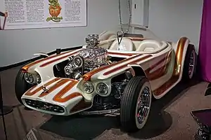 A white car with brown stripes, with open wheels and a clear bubble canopy over twin seats, and exposed, chromed engine with a blower.  Beatnik Bandit II and a few of Roth's other cars are also on display in this museum.