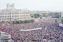 A crowd of thousands stands in a plaza. Many carry large banners.