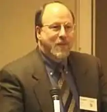 A middle-aged man with balding blond hair, a white beard, and glasses stands beside a microphone in his grey suit and pattern tie.