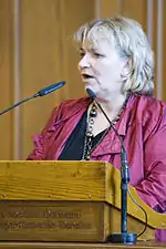 Head and upper body of a plump middle-aged woman with grey-blonde hair speaking at a wooden lectern, two slim microphones in front of her.