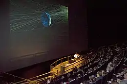 A film theater with a giant screen. A man is talking at a stage in front of audiences.