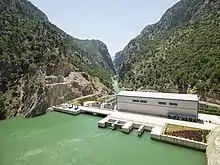 Green water behind a dam with a building on top, in a small steep-sided valley