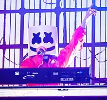 Marshmello holding a microphone in his right hand with his left hand raised up
