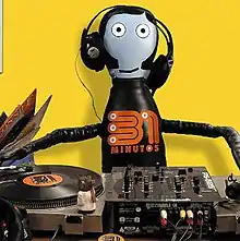 A bowling pin-shaped puppet dressed in black with an orange 31 minutos logo in his chest is behind a turntable with a 31 minutos LP placed on it, behind the puppet theres a group of LPs; the puppet is wearing DJ headphones, has blue skin, round white eyes black hair and sideburns in a yellow background.