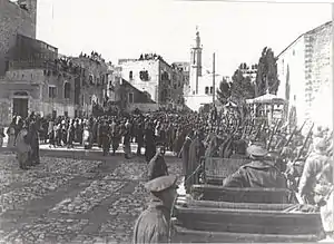 City buildings, some with people looking down from roofs, people and soldiers in a large square, with motor car in foreground