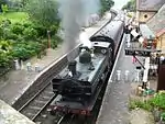 A green pannier tank locomotive is seen from above and to the front as it waits at a station with two railway tracks and platform. The platform on the left includes a small wooden shelter, benches, lamps, flowers, and, near the end of the platform, three milk churns positioning across the platform to prevent people passing. The platform on the right, where the train is waiting, includes a brick building, more flowers, and again, three milk churns arranged to stop people passing. There is also a signal near the locomotive, and its black and white semaphore arm is pointing downward. The train includes seven coaches in maroon. A plume of grey smoke comes from the locomotive chimney, and a smaller plume of white steam from the safety valve near the cab.