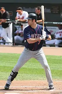 A man in a left-handed batting stance wearing pinstriped gray pants, a black shinguard on his right leg, a dark blue baseball jersey, and a dark-colored batting helmet.