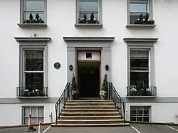 A flight of stone steps leads from an asphalt car park up to the main entrance of a white two-story building. The ground floor has two sash windows, the first floor has three shorter sash windows. Two more windows are visible at basement level. The decorative stonework around the doors and windows is painted grey.