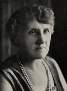 An older white woman with coiffed grey hair, wearing a dress or top with a low square neckline, and two strands of pearls