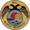 A dark-yellow circular seal with a smaller circle inside it that portrays a fortified tower and wall behind blue waves. The smaller circle is enclosed by a black two-headed bird with the text T.C. above and Alanya Belediyesi below.