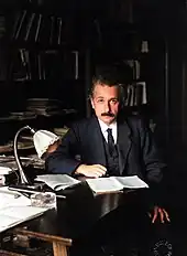 Einstein, sitting at a table, looks up from the papers he is reading and into the camera.