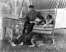 A man sits on a swing, writing on a crude desk. Two dogs, Rough Collies, sit on his left. Another lies on the ground at his feet.