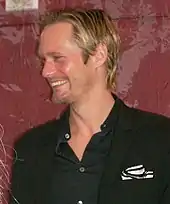 A man with blond hair and dressed in a black shirt and blazer smiles to his right