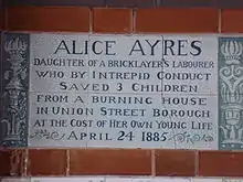 A tablet formed of six standard sized tiles, bordered by green flowers in the style of the Arts and Crafts movement, with an inscription in green capital letters on a white background