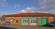 A large, sunlit, single-storey orange brick building with a roof covered with red tiles. On the left are two small green doors, in the centre are three large, green, shutter doors. Atop the roof is an aerial.