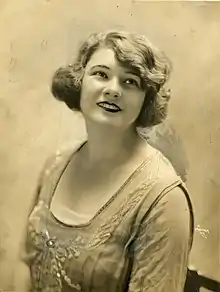 A sepia-toned photograph of a young white woman, smiling; her wavy hair is bobbed; she is wearing an embroidered dress with a low square neckline.