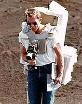 Man around age 40 with sunglasses and a large backpack takes a photograph with a camera mounted on his chest