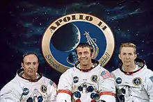 Mitchall, a smiling Shepard and Roosa wearing their Apollo space suits without the helmets. Their suits sport their names, the mission patch on the left breast and NASA patch on the right. The wear the American flag on their left sleeve. Shepard has red rings in his arms. In the background is a giant mission patch, surrounded by black space and stars.