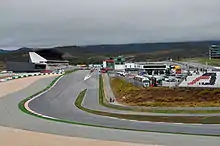 A tarmac course in hilly terrain, lined with concrete and gravel run-off areas, passes between a large grandstand and the pit garages.