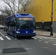 A front view of a curved mostly blue bus passing a bright yellow building