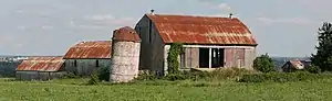 A short silo in the centre of the photograph is slanted slightly to the right, topped by a conical red roof. Three barns form a V shape behind the silo. To its right is a large barn, with slanted red roof and open doors. Perpendicular to it are two similar but smaller barns in series, visible to the left of the silo.