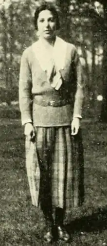 A young woman with dark hair, standing outdoors; she is wearing a blouse with a white collar under a belted sweater, with a pleated checked skirt