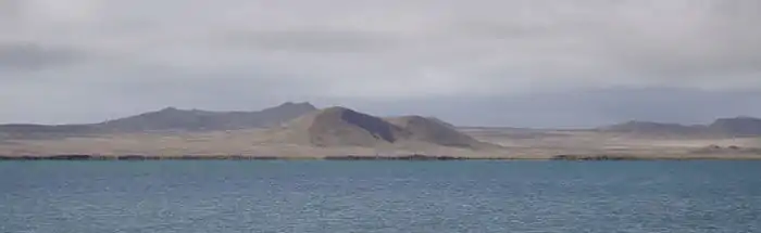 Smooth, rounded hills and flatlands covered in golden-brown vegetation lie beyond a lake under heavy cloud.