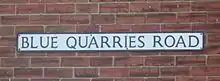On a red brick wall, a rectangular sign bears the name "Blue Quarries Road" in black, upper case letters on a white background.