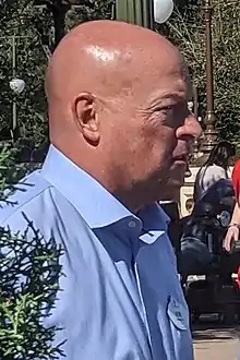 Man in a blue collared suit partly behind some shrubbery