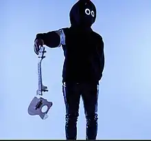 BoyWithUke, wearing his mask with two white circle lights for eyes and a black hoodie and pants, holds a broken ukulele in front of himself while looking away
