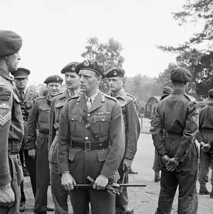 General officer with gloves and cane in hand, talking to a sergeant, surrounded by other officers and men