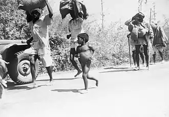 A small, naked, pot-bellied boy runs beside a line of men carrying large bundles on their heads. Some of the men are also running. All are on a road. A military vehicle is partially visible beside them.