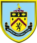 A shield-shaped crest with a mainly light blue background. The crest features a stork at the top, standing on hills and cotton plants. Further down, a black band including a hand and two bees; a wavy, claret-coloured line, and a lion. "Burnley Football Club" is written at the bottom.