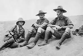 An informal black and white group portrait of three men in military uniform. They are sitting on the ground in what appears to be a desert.