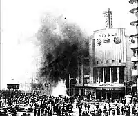 A crowd of bystanders stands outside a burning building, with black smoke coming out of the windows.