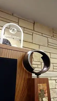 A wooden trophy with a metal oval on top standing in front of a podium, which has a plexiglass trophy on top