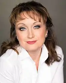 A close-up posed upper body shot of a woman. Her head is tilted to her left. She has short, black hair and pale blue eyes. She wears a grey/white fleecy boa/scarf over her black top. She has make up and red lipstick and sports an earring in her left ear (right ear is not visible).
