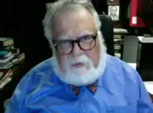 Celal Şengör is a white caucasian male, he has white hair and beard, he is chubby, he has full beard and slightly receeding hairline and he has glasses.