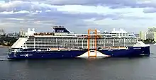 A blue-painted cruise ship is viewed from her starboard (right) side as she sails out of port; along the middle of the vessel is an orange sheltered platform that moves on a track on the starboard side
