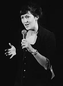 Black-and-white photo of Caucasian woman standing onstage, speaking into a microphone