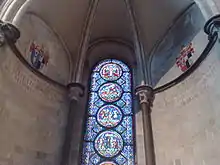A semi-circular area with stone walls, a domed ceiling and a tall stained glass window with prominent circular decorations.