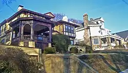 Two houses, one in a Tudor Revival style with a garage built into the ground beneath it and the other of wood with a stone foundation, seen from a street slopping downhill from them.
