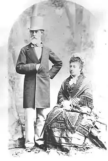 An old man in a top hat with a white beard stands next to a sitting woman in a decorative dress