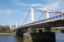 Suspension bridge crossing a wide river, under blue skies with clouds. The main cables are red and the towers white.