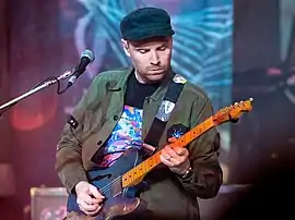 A man wearing a dark cap and a green jacket plays the guitar